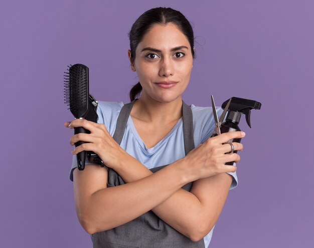 Junge schöne Frau Friseur in Schürze halten Sprühflasche Trimmer Maschine und Haarbürste nach vorne mit selbstbewussten Ausdruck über lila Wand stehen