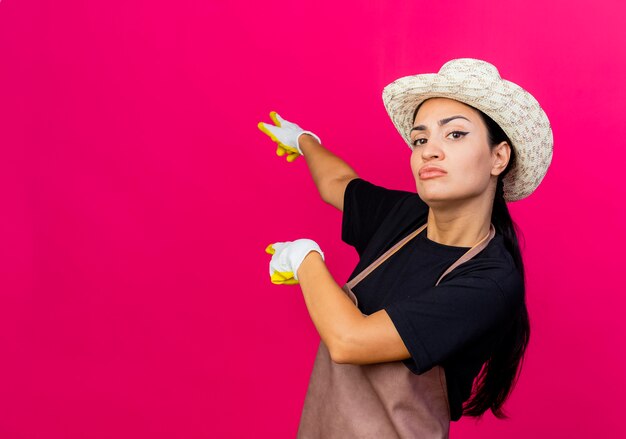 Junge schöne Frau Gärtner in Gummihandschuh Schürze und Hut zeigt mit Zeigefinger zurück suchen mit ernstem Gesicht über rosa Wand stehen