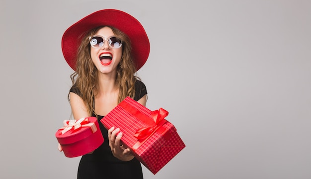 Junge schöne Frau hält Geschenke, schwarzes Kleid, roter Hut, Sonnenbrille, glücklich, lächelnd, sexy, elegant, Geschenkboxen, feiern, positiv, emotional