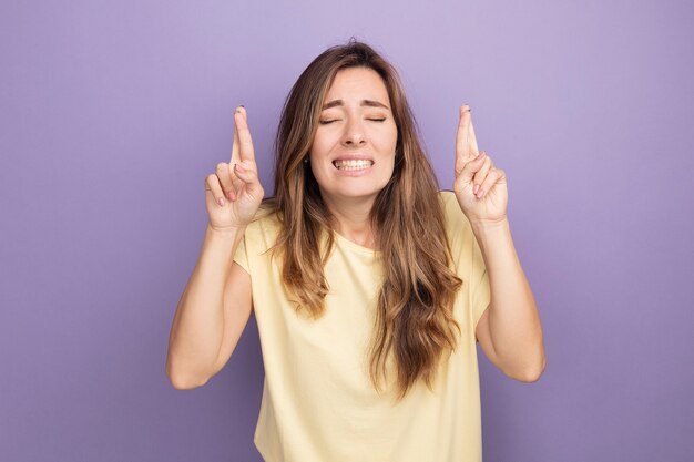 Junge schöne Frau im beigen T-Shirt, die mit geschlossenen Augen einen wünschenswerten Wunsch mit Hoffnungsausdruck macht