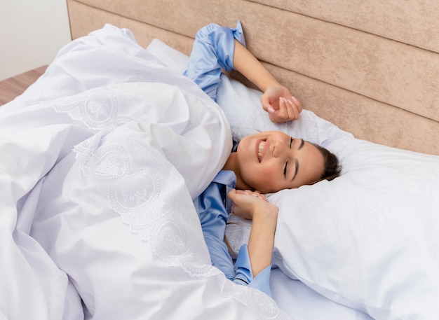 Junge schöne Frau im blauen Pyjama, der auf Bett liegt, das auf weichen Kissen ruht, die aufwachen, die ihre Hände im Schlafzimmerinnenraum auf hellem Hintergrund strecken