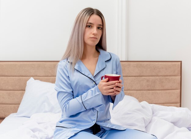 Junge schöne Frau im blauen Pyjama, der auf Bett mit Tasse Kaffee sitzt und Kamera mit ernstem Gesicht im Schlafzimmerinnenraum auf hellem Hintergrund betrachtet
