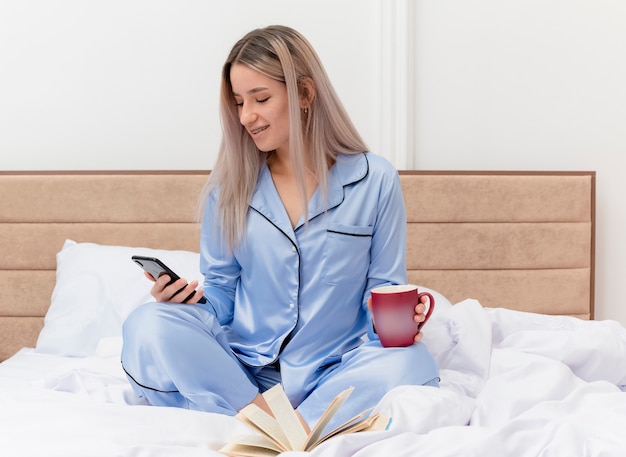 Junge schöne Frau im blauen Pyjama, der auf Bett mit Tasse Kaffee unter Verwendung des Smartphones sitzt, das im Schlafzimmerinnenraum auf hellem Hintergrund lächelt