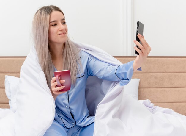 Junge schöne Frau im blauen Pyjama, der auf Bett unter Blenket mit Tasse Kaffee tut, der selfie unter Verwendung des Smartphones glücklich und positiv im Schlafzimmerinnenraum auf hellem Hintergrund tut