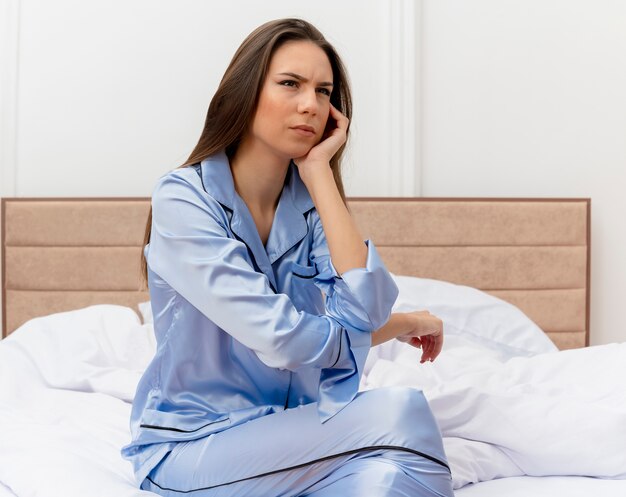 Junge schöne Frau im blauen Pyjama, der auf Bettblick mit ernstem Stirnrunzeln im Schlafzimmerinnenraum auf hellem Hintergrund sitzt