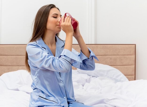 Junge schöne Frau im blauen Pyjama, der auf dem Bett sitzt, das Kaffee glücklich und positiv im Schlafzimmerinnenraum auf hellem Hintergrund trinkt