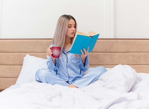 Junge schöne Frau im blauen Pyjama, die auf dem Bett mit einer Tasse Kaffee sitzt und ein Buch im Schlafzimmer liest