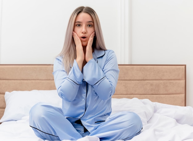 Junge schöne Frau im blauen Pyjama, die auf dem Bett sitzt und in die Kamera schaut, erstaunt und überrascht im Schlafzimmerinnenraum