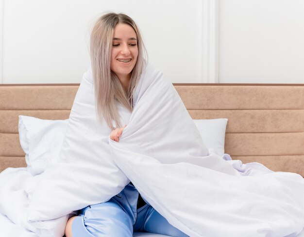 Junge schöne Frau im blauen Pyjama, die auf dem Bett sitzt und in eine Decke einwickelt, die glücklich und positiv im Schlafzimmerinnenraum lächelt