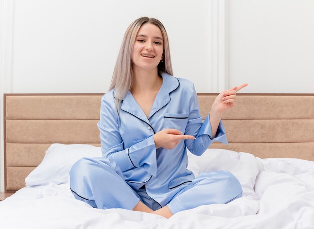 Junge schöne Frau im blauen Pyjama, die auf dem Bett sitzt und mit den Zeigefingern zur Seite zeigt, die fröhlich im Schlafzimmerinnenraum lächelt