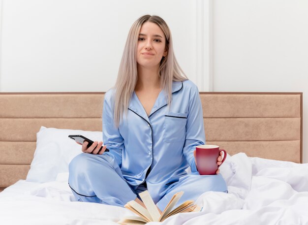 Junge schöne Frau im blauen Pyjama sitzt auf dem Bett mit einer Tasse Kaffee mit Smartphone und lächelt im Schlafzimmer