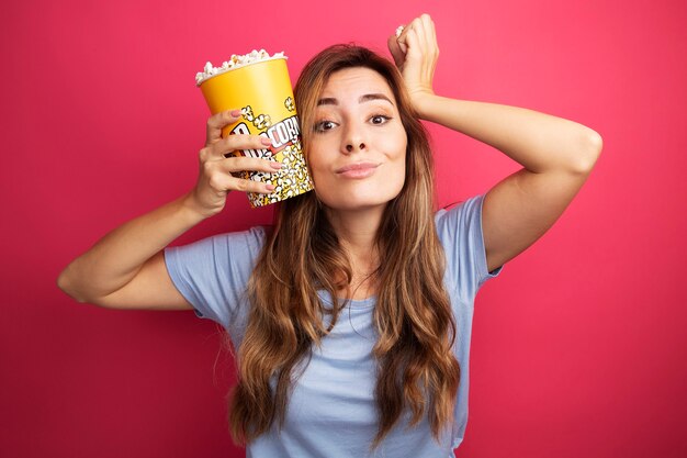 Junge schöne Frau im blauen T-Shirt, die Eimer mit Popcorn hält und die Kamera glücklich und fröhlich lächelt, die über rosafarbenem Hintergrund steht