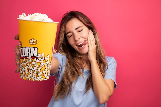 Junge schöne Frau im blauen T-Shirt, die Eimer mit Popcorn-hapy und positiver Zunge hält