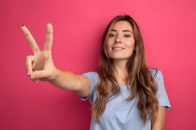 Junge schöne Frau im blauen T-Shirt, die mit einem Lächeln auf dem Gesicht beiseite schaut, das ein V-Zeichen auf rosafarbenem Hintergrund zeigt