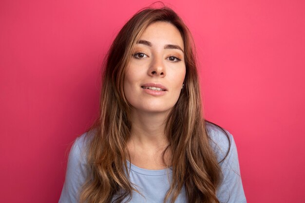 Junge schöne Frau im blauen T-Shirt mit Blick in die Kamera lächelt zuversichtlich stehend über rosa Hintergrund