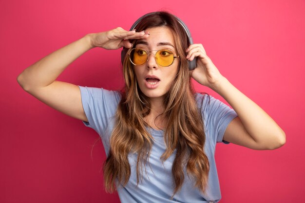 Junge schöne Frau im blauen T-Shirt mit gelber Brille mit Kopfhörern, die mit der Hand über dem Kopf weit weg schauen