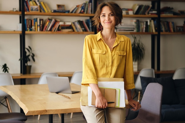 Junge schöne Frau im gelben Hemd, die sich mit Notizblock und Papieren in der Hand auf den Schreibtisch lehnt, während sie im modernen Büro glücklich in die Kamera blickt