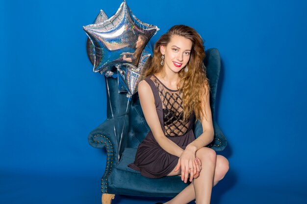 Junge schöne Frau im grauen Kleid, das auf einem blauen Sessel mit silbernen Luftballons sitzt