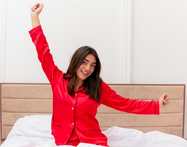 Junge schöne Frau im roten Pyjama, die im Bett sitzt und sich ausdehnt und glücklich und positiv lächelt im Schlafzimmerinnenraum aufwacht