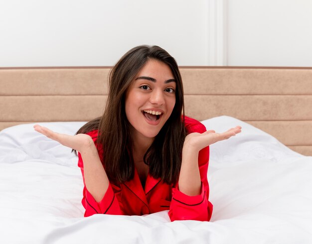 Junge schöne Frau im roten Pyjama, die sich auf dem Bett entspannt und mit glücklichem Gesicht im Schlafzimmerhintergrund lächelt