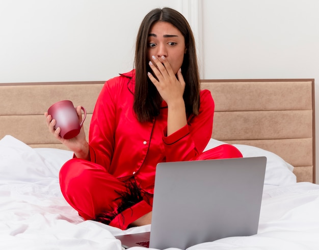 Junge schöne Frau im roten Schlafanzug, der auf Bett mit Laptop und Tasse Kaffee sitzt, der im Schlafzimmerinnenraum auf hellem Hintergrund erstaunt und überrascht schaut