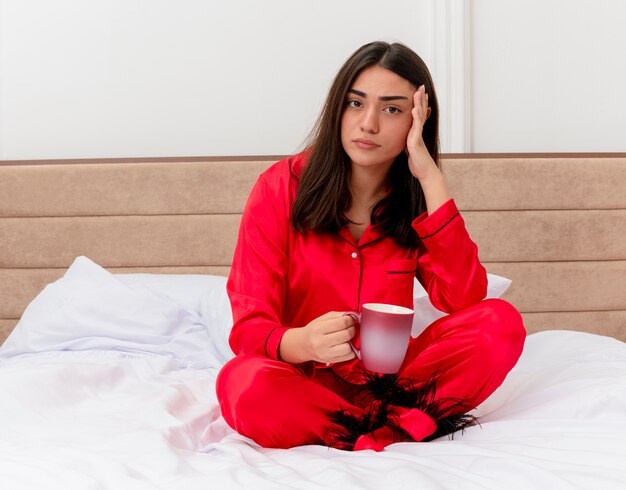 Junge schöne Frau im roten Schlafanzug, der auf Bett mit Tasse Kaffee betrachtet Kamera betrachtet, die im Schlafzimmerinnenraum auf hellem Hintergrund verwirrt wird