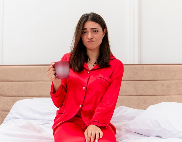 Junge schöne Frau im roten Schlafanzug, der auf Bett mit Tasse Kaffee betrachtet Kamera betrachtet verwirrt und unzufrieden in Schlafzimmerinnenraum auf hellem Hintergrund
