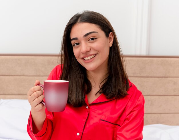 Junge schöne Frau im roten Schlafanzug, der auf Bett mit Tasse Kaffee sitzt und Kamera betrachtet, die fröhlich im Schlafzimmerinnenraum auf hellem Hintergrund lächelt