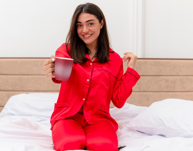 Junge schöne Frau im roten Schlafanzug, der auf Bett mit Tasse Kaffee sitzt und Kamera betrachtet, die fröhlich im Schlafzimmerinnenraum auf hellem Hintergrund lächelt