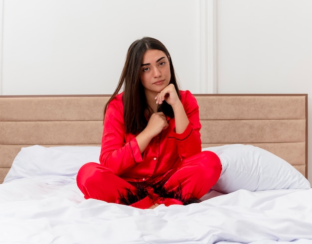 Junge schöne Frau im roten Schlafanzug, der auf Bett sitzt und Kamera mit ernstem Gesicht im Schlafzimmerinnenraum auf hellem Hintergrund betrachtet