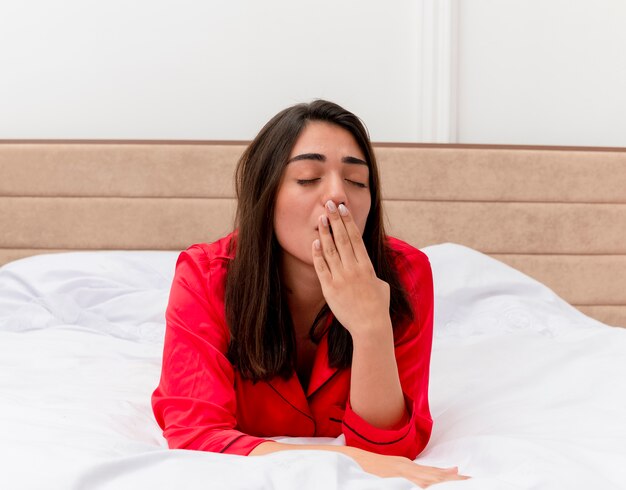 Junge schöne Frau im roten Schlafanzug, der auf dem Bett liegt, das sich entspannt, will gähnend im Hauptinnenraum auf hellem Hintergrund schlafen