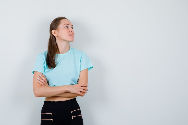 Junge schöne Frau im T-Shirt, Hose mit verschränkten Armen, wegschauen und fröhlich aussehend, Vorderansicht.