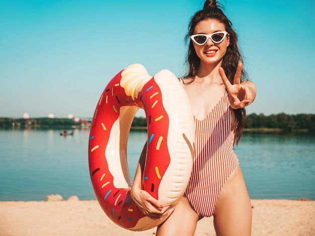 Junge schöne Frau in Badebekleidung und Sonnenbrille, die am Strand mit aufblasbarem Donut aufwirft