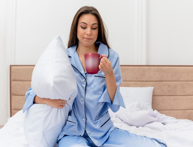 Junge schöne Frau in den blauen Pyjamas, die auf Bett mit Kissen und Tasse Kaffee aufwachen und Morgenmüdigkeit im Schlafzimmerinnenraum auf hellem Hintergrund aufwachen