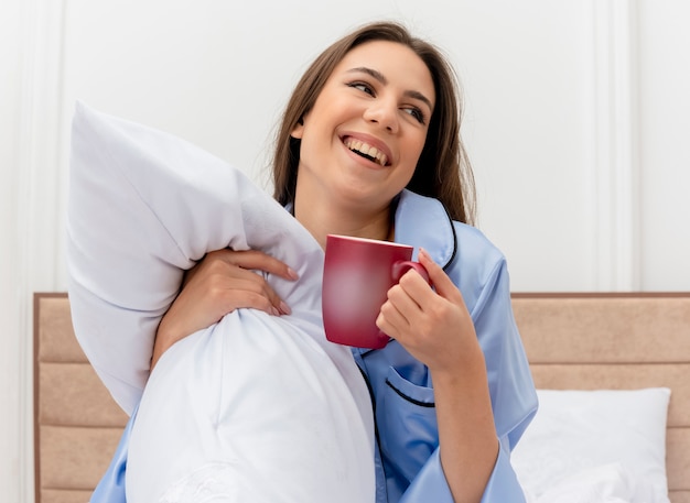 Junge schöne frau in den blauen pyjamas, die auf bett mit kissen und tasse kaffee sitzen und beiseite lächelnd im schlafzimmerinnenraum auf hellem hintergrund schauen