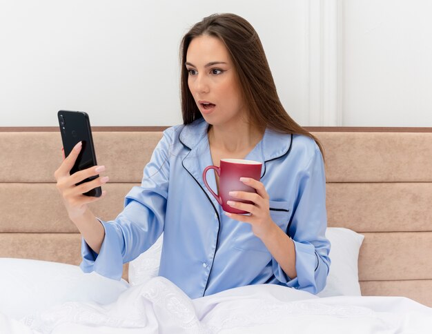 Junge schöne Frau in den blauen Pyjamas, die auf Bett mit Tasse Kaffee sitzen und ihren Smartphonebildschirm betrachten, der im Schlafzimmerinnenraum auf hellem Hintergrund erstaunt und überrascht wird