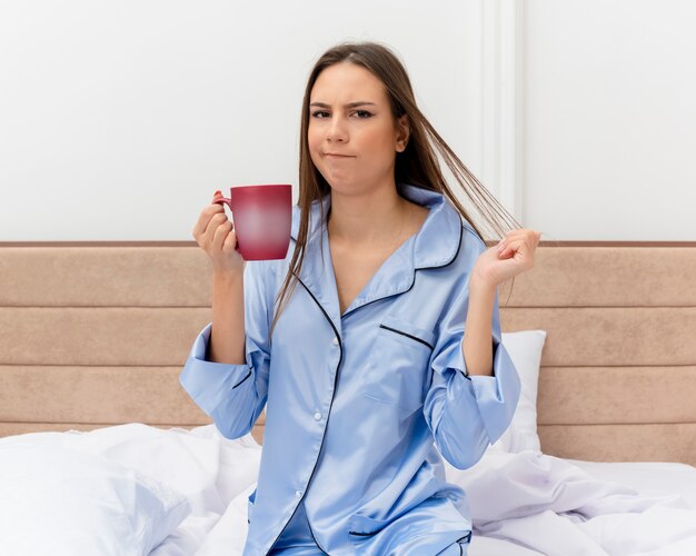 Junge schöne Frau in den blauen Pyjamas, die auf Bett mit Tasse Kaffee suchen, der an der Kamera unzufrieden ist, die im Schlafzimmerinnenraum auf hellem Hintergrund aufwacht