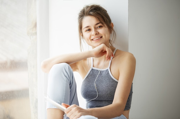 Junge schöne Frau in den lächelnden Kopfhörertafeln, die das Streamen von Musik hören, die auf Boden über weißer Wand sitzt.