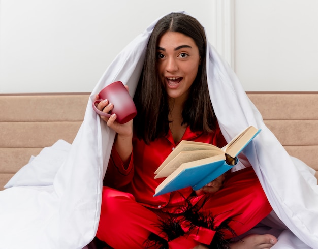 Junge schöne Frau in den roten Pyjamas, die auf Bett sitzen, das in Decke mit Tasse Kaffee und Buch glücklich und positiv lächelnd im Schlafzimmerinnenraum auf hellem Hintergrund wickelt