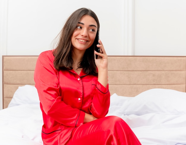 Junge schöne Frau in den roten Pyjamas, die auf dem Bett lächelnd lächeln, während auf Handy im Schlafzimmerinnenraum auf hellem Hintergrund sprechen