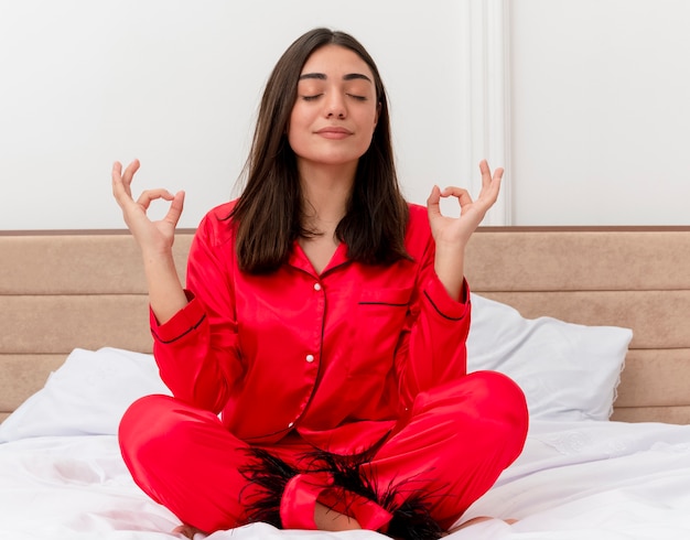 Junge schöne frau in den roten pyjamas, die auf dem entspannenden bett sitzen und meditationsgeste mit den fingern mit den geschlossenen augen im schlafzimmerinnenraum auf hellem hintergrund machen
