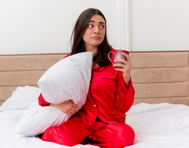 Junge schöne frau in den roten pyjamas, die im bett mit kissen halten, das eine tasse hält, die beiseite schaut, verwirrt im schlafzimmerinnenraum auf hellem hintergrund