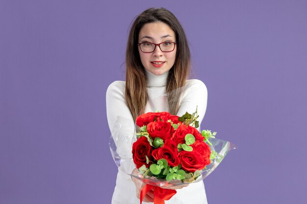 junge schöne Frau in der Freizeitkleidung, die Strauß der roten Rosen hält, die Kamera glücklich und positivmiling fröhlich Valentinstagkonzept betrachten stehen über lila Wand