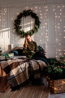 Junge schöne frau in einem pullover in einem weihnachtlich dekorierten raum sitzt auf dem hintergrund eines weihnachtsbaumes