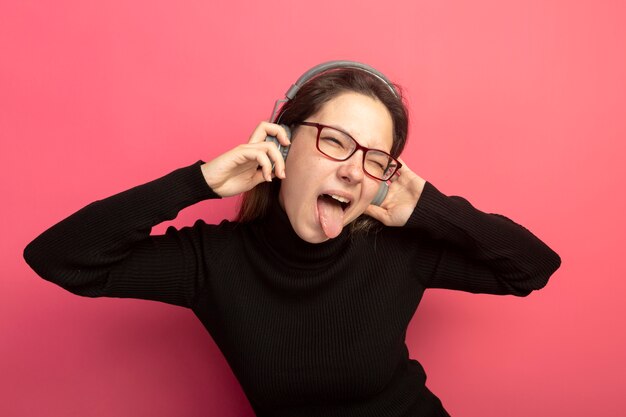 Junge schöne Frau in einem schwarzen Rollkragenpullover und Brille mit Kopfhörern glücklich und aufgeregt herausstehende Zunge, die über rosa Wand steht