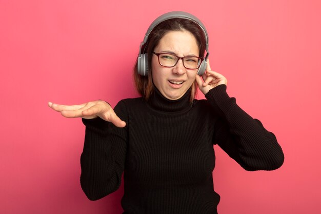 Junge schöne Frau in einem schwarzen Rollkragenpullover und einer Brille mit Kopfhörern, die ihre Lieblingsmusik genießen, die über rosa Wand steht