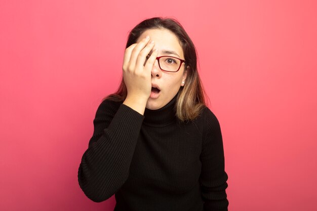 Junge schöne Frau in einem schwarzen Rollkragenpullover und in den Brillen, die an der Kamera verwirrt sind, die ein Auge mit der Hand bedeckt bedeckt, die über rosa Wand steht