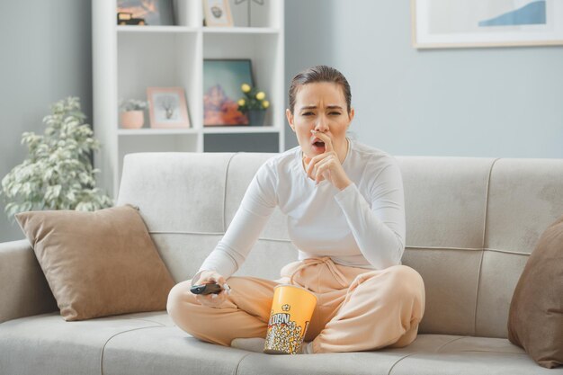 Junge schöne Frau in Freizeitkleidung, die auf einer Couch zu Hause sitzt, mit einem Eimer Popcorn, der die Fernbedienung hält und in die Kamera schaut, verwirrt versucht, sich an etwas Wichtiges zu erinnern