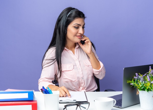 Junge schöne Frau in Freizeitkleidung, die ein Headset mit Mikrofon trägt und selbstbewusst lächelnd am Tisch mit Laptop über blauem Hintergrund sitzt und im Büro arbeitet