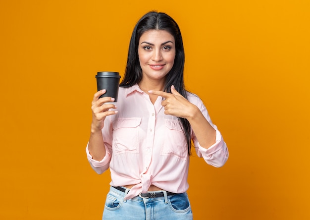 Junge schöne Frau in Freizeitkleidung, die eine Kaffeetasse hält und mit dem Zeigefinger darauf zeigt, lächelt selbstbewusst über orangefarbener Wand